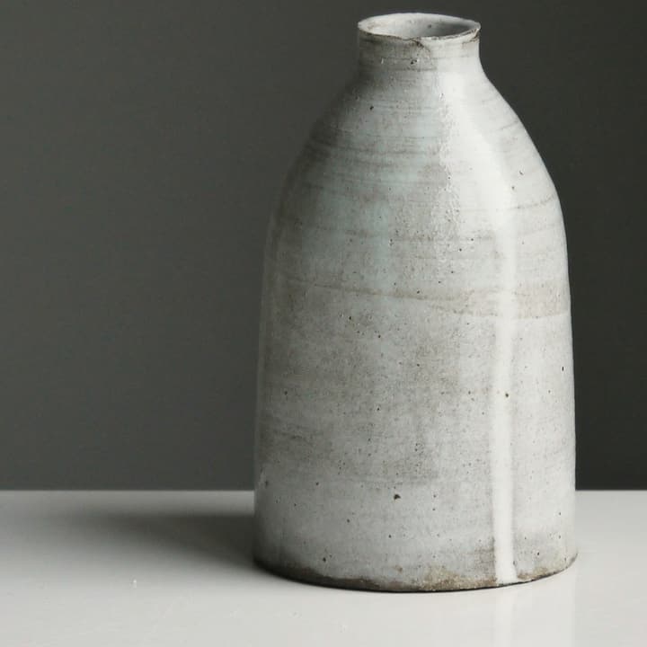 Grey speckled bottle vase on a white shelf against a dark wall, detail of the glaze