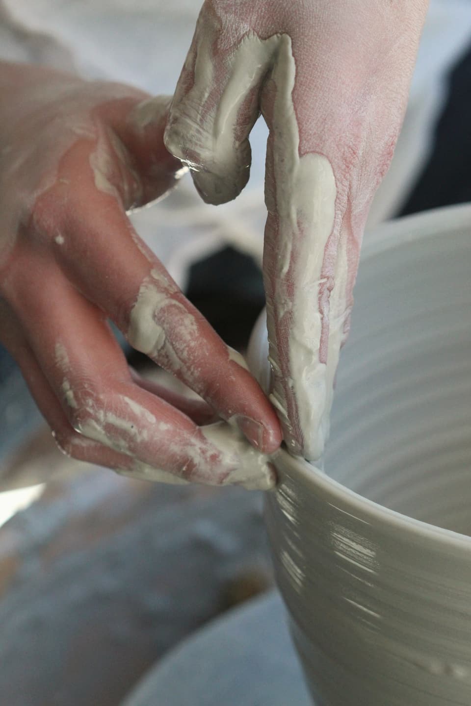 Hands covered in wet grey clay shaping the rim of a bowl